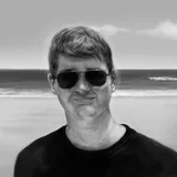 Schwarzweiß-Porträt: Mann mit Sonnenbrille lächelt am Strand vor Meer und Wolkenhimmel.