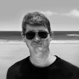 Grayscale portrait of smiling man in sunglasses and black t-shirt on beach with ocean waves.