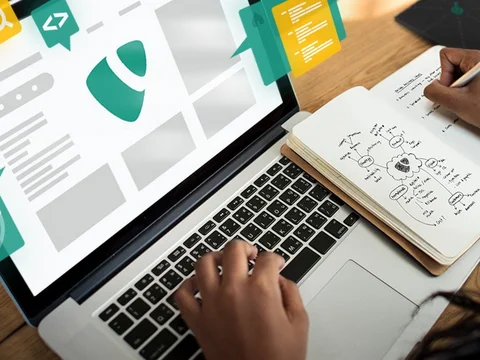 Person schreibt Notizen in ein Notizbuch neben einem Laptop mit grafischen Elementen auf dem Bildschirm.