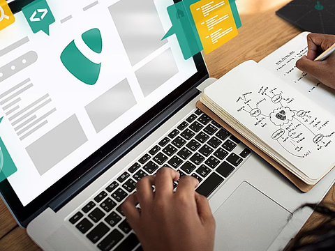 Person schreibt Notizen in ein Notizbuch neben einem Laptop mit grafischen Elementen auf dem Bildschirm.