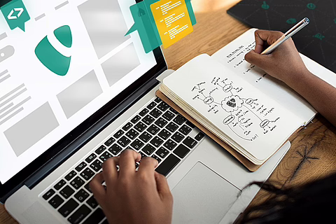 Over-shoulder view of Black woman at desk using laptop with teal/yellow e-commerce UI, writing notes.