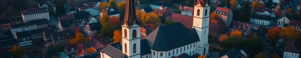 Luftaufnahme einer europäischen Kleinstadt mit zwei Kirchtürmen, roten Dächern, Herbstbäumen und Fluss im Hintergrund.