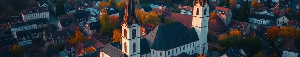 Luftaufnahme einer europäischen Kleinstadt mit zwei Kirchtürmen, roten Dächern, Herbstbäumen und Fluss im Hintergrund.