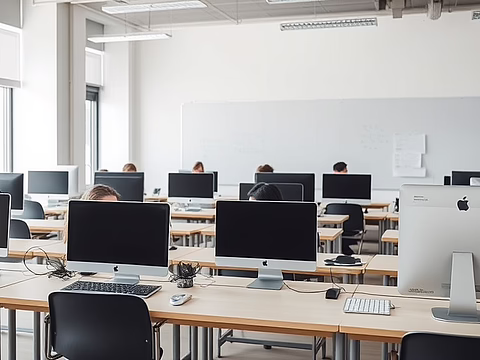 Klassenzimmer mit vielen iMac-Computern auf Tischen; Studierende arbeiten, große Fenster links, Whiteboard an der Wand.
